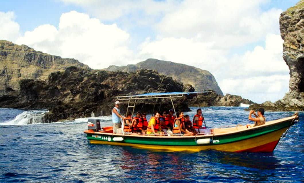 A group of people wearing life jackets sit on a colorful motorboat in the blue ocean off Easter Island, surrounded by Rapa Nui’s rocky cliffs under a partly cloudy sky.