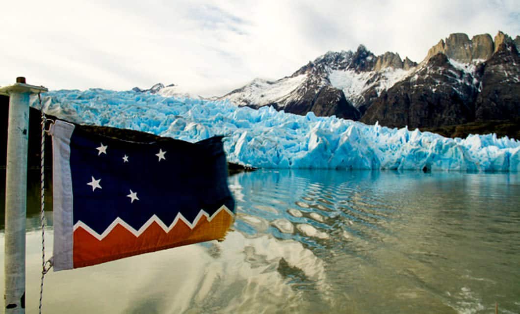A flag with blue, orange, and white zigzag stripes and white stars flutters on a boat near icy blue Patagonian glaciers and rugged mountains reflected in the water under a cloudy sky, not far from a cozy Eco-Camp lodge.