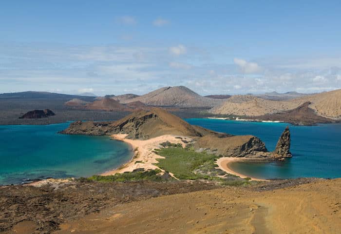 A scenic view of Bartolomé Island in the Galapagos Islands, showing turquoise waters, sandy beaches, rugged volcanic terrain, and the distinctive Pinnacle Rock jutting from the shoreline under a partly cloudy sky.