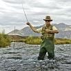 A person wearing waders and a hat is fly fishing in a river, holding a fishing rod with mountains and cloudy skies in the background, likely benefiting from great guides on where to cast.