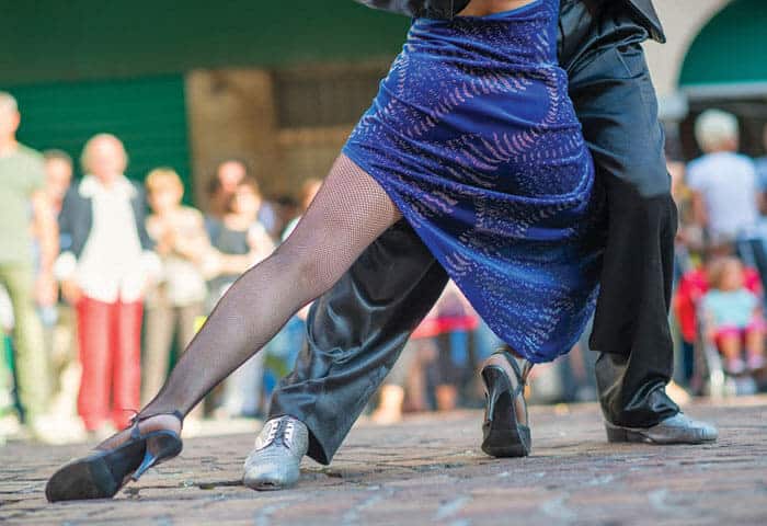 Two dancers perform a passionate tango on a cobblestone street—one of the classic things to do in Argentina. The woman wears a blue dress and fishnet stockings, the man wears black trousers and silver shoes. A blurred crowd watches in the background.