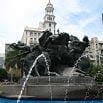 A large fountain with dynamic bronze sculptures and water jets in a city square, framed by tall historic buildings and a clock tower under a partly cloudy sky—a true bucket list spot for urban explorers.