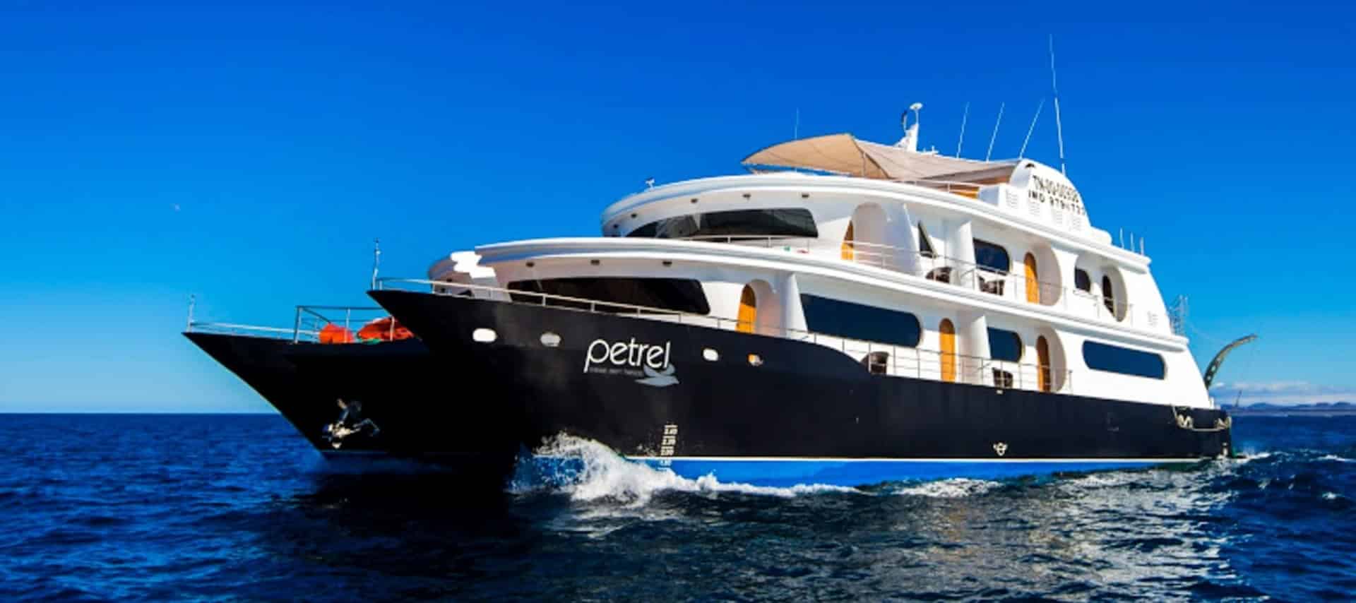 A large, modern yacht named Petrel cruises on a calm blue ocean under a clear sky, offering multiple decks and outdoor seating—perfect for unforgettable Galapagos cruises around the stunning Galapagos Islands.
