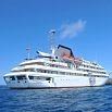 A large white cruise ship is sailing on calm blue water under a clear sky. With multiple decks and glass windows, the ship offers excellent service as people enjoy the view from the top deck.