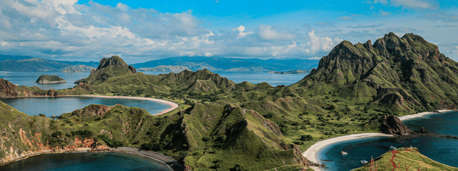 A panoramic view of lush green hills and curving coastlines surrounding clear blue bays under a bright sky with scattered clouds, featuring rugged mountains in the background—an iconic scene from any Galapagos Islands travel guide.