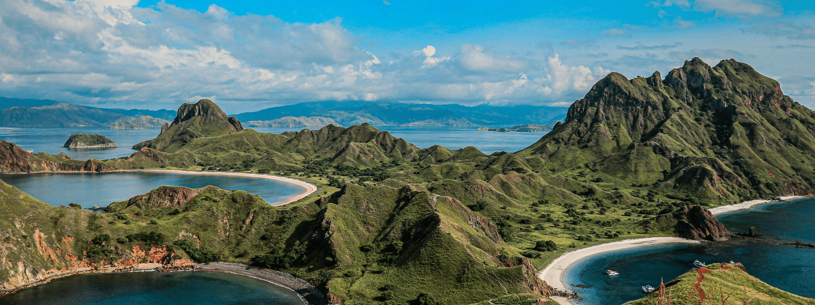 A panoramic view of lush green hills and curving coastlines surrounding clear blue bays under a bright sky with scattered clouds, featuring rugged mountains in the background—an iconic scene from any Galapagos Islands travel guide.