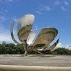 A terrific large metallic flower sculpture with open petals stands in an outdoor plaza, surrounded by greenery and under a bright blue sky with scattered clouds—a perfect stop on your trip.