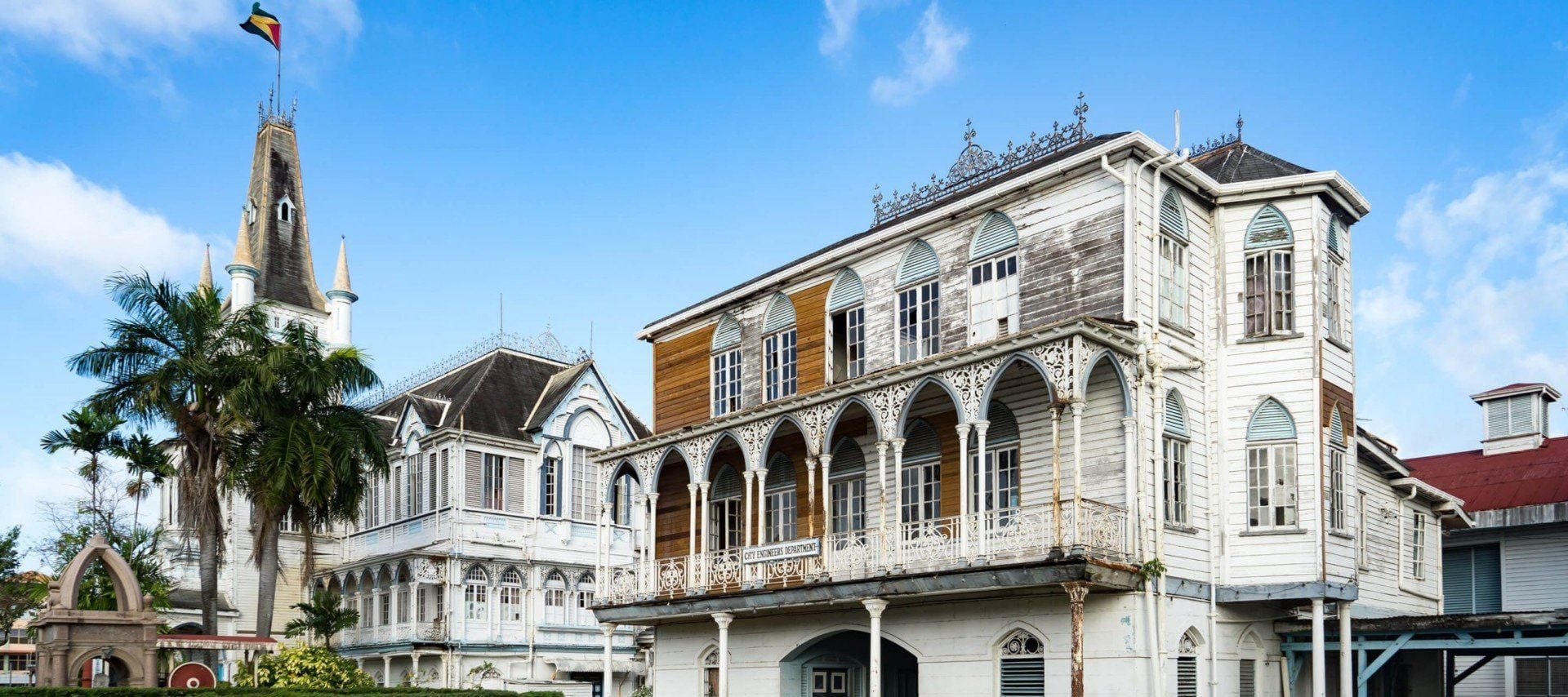 A large, ornate wooden colonial-style building with arched windows and decorative trim stands beside an old clock tower and palm trees under a bright blue sky—a perfect scene to welcome travelers arriving on flights to Guianas.