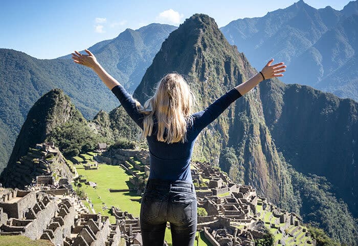 A person with long blond hair stands with arms raised, overlooking the ancient stone ruins of Machu Picchu in South America, surrounded by green mountains under a clear blue sky.
