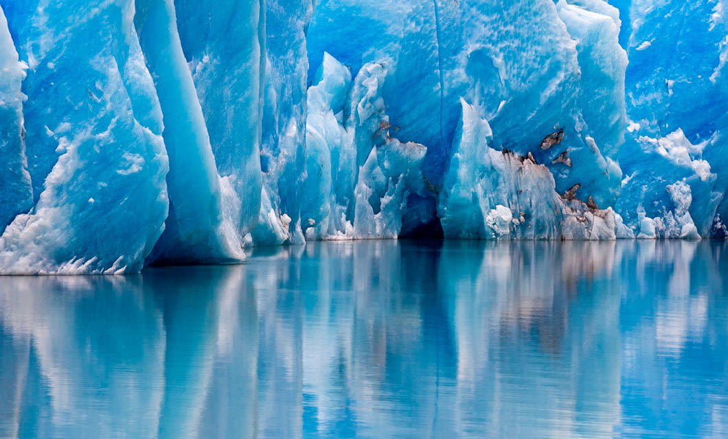Massive blue glaciers reflected in calm, clear water, with jagged ice formations and smooth surfaces creating a striking, cold landscape near the serene Tierra Patagonia hotel.