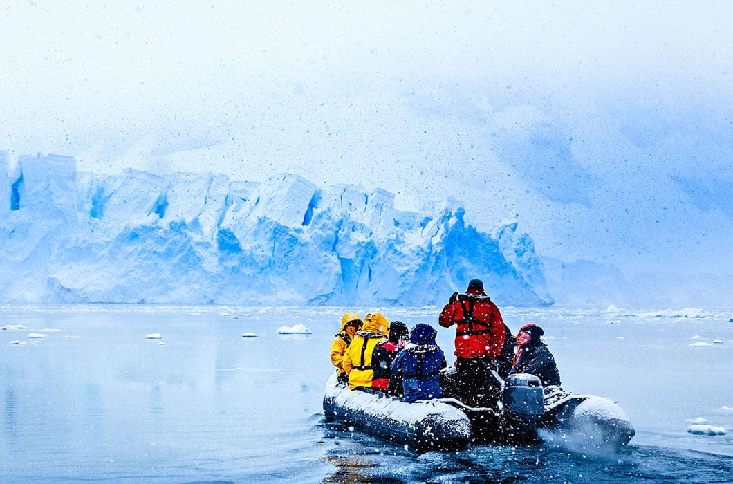 A group of people in colorful jackets ride an inflatable boat on icy water, approaching a massive glacier under snowfall—one of the unforgettable Antarctica activities with floating ice all around.