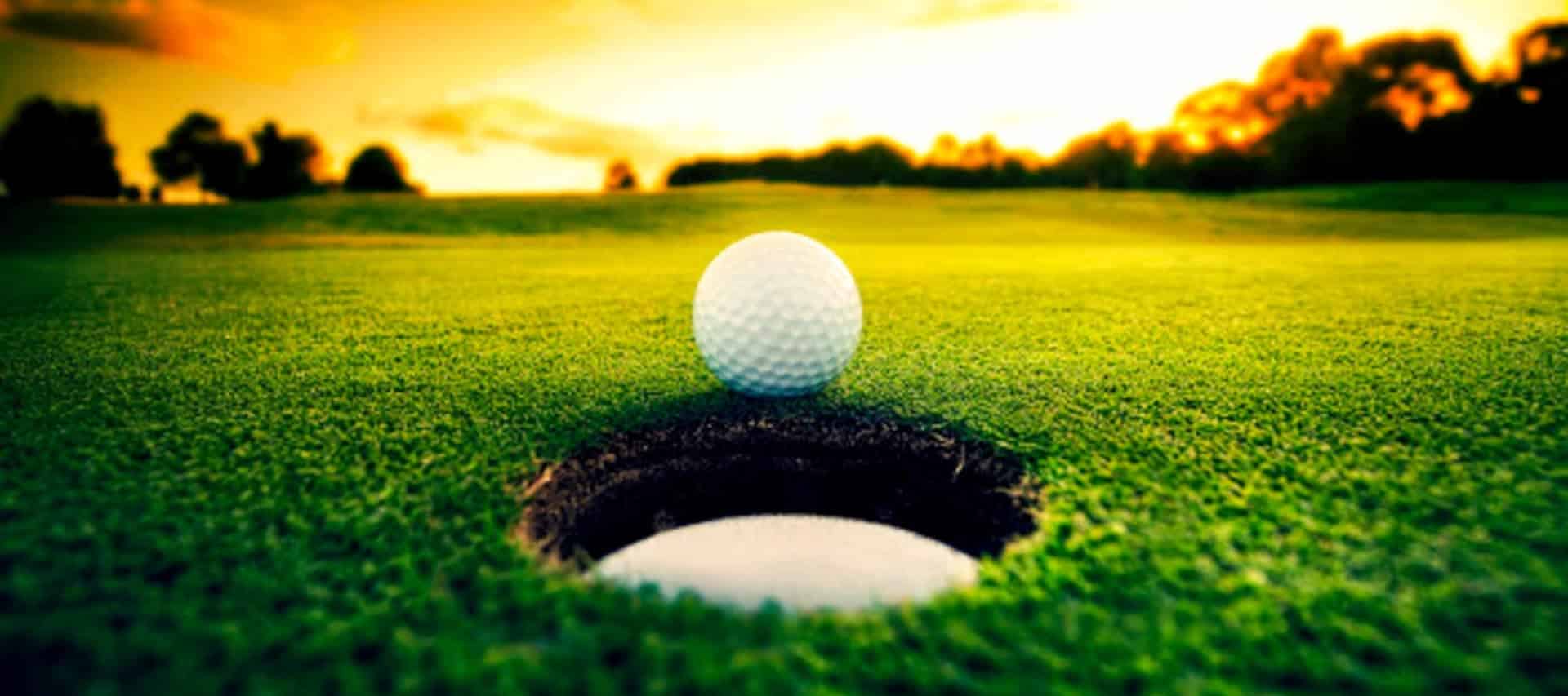 A close-up of a golf ball about to fall into a hole on a green golf course at sunset, with trees silhouetted on the horizon—perfect inspiration for unforgettable Peru golf tours.