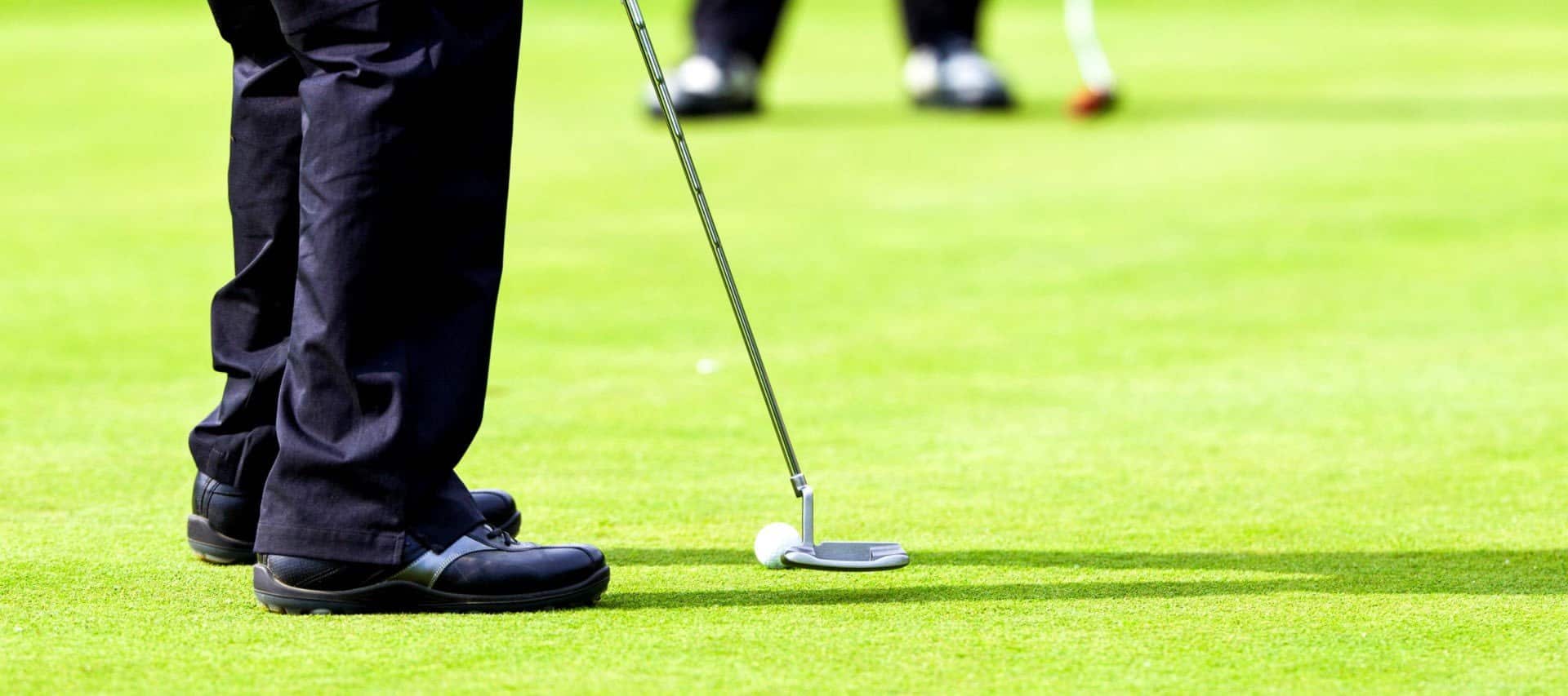 A close-up of a golfer’s legs and shoes as they prepare to putt on a green during one of the Chile Golf Tours, with a golf ball and putter in position. Another golfer is visible in the blurred background.