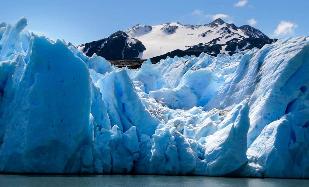 Jagged blue icebergs and glaciers rise from the water, with snow-capped mountains in the background under a partly cloudy sky—an awe-inspiring view best enjoyed from a luxury lodge like Awasi Patagonia, an exceptional Patagonia hotel.