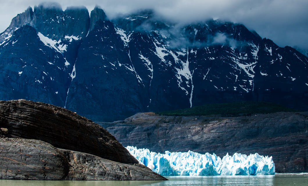 A bright blue glacier sits at the base of dark, rugged mountains with patches of snow, under a cloudy sky. Near calm, greenish water and a rocky outcrop, this stunning scene captures the essence of Torres del Paine in Patagonia.