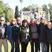 A group of people stand outdoors in a line, smiling at the camera during their trip. Trees and a large building are visible in the background on a sunny day.