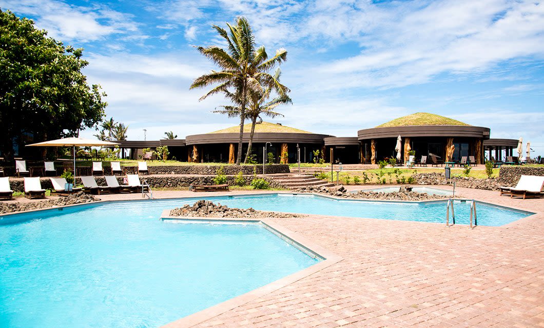 A luxurious eco village resort with two outdoor swimming pools, surrounded by lounge chairs and palm trees, overlooks a modern, round building in the background under a bright blue Easter Island sky.
