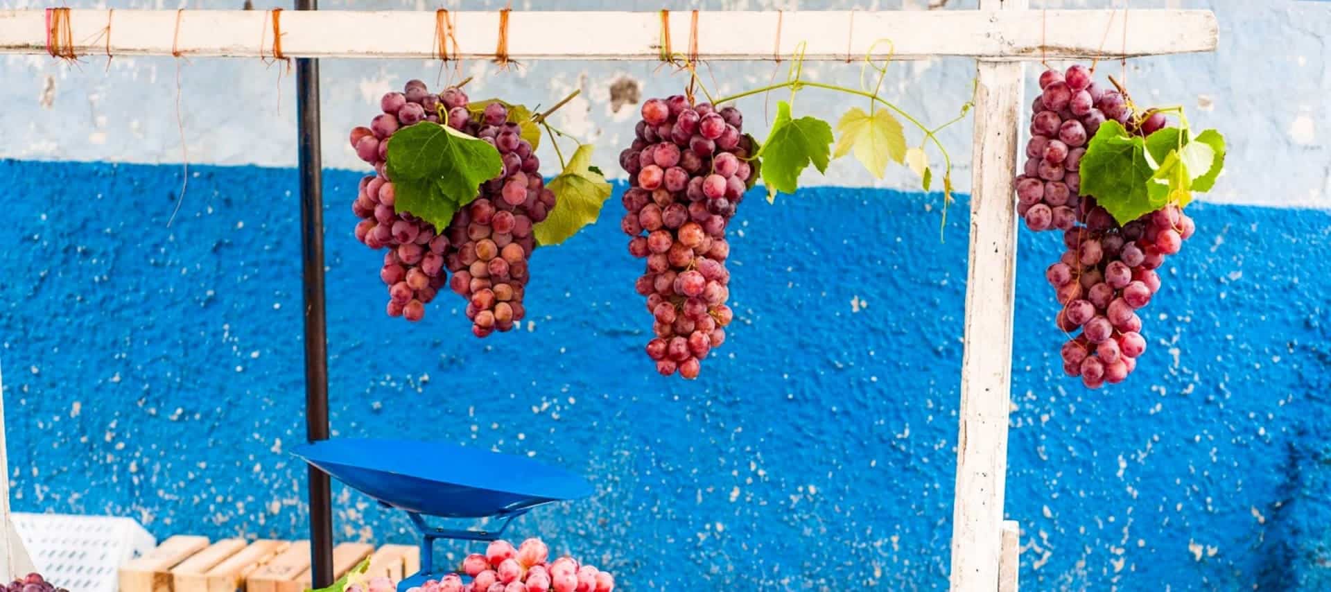 Four large clusters of red grapes hang from a wooden beam in front of a blue and white textured wall; a blue weighing scale and more grapes sit below, evoking the charm of Bolivia Wine Tours.