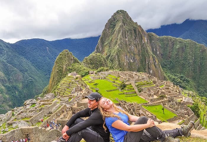 Two people sit back-to-back, smiling, on a rocky ledge overlooking the ancient ruins of Machu Picchu in Peru—a must-see for travel lovers—surrounded by lush green terraces and a mountain peak under cloudy skies.