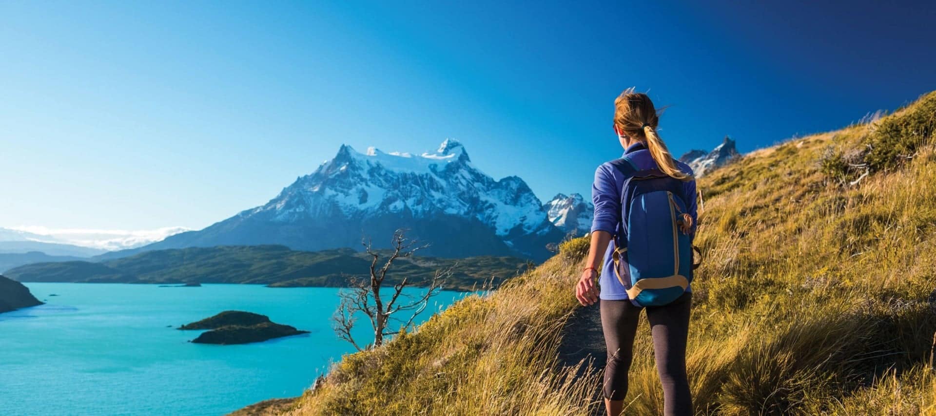 A woman with a blue backpack hikes along a grassy trail overlooking a turquoise lake and snow-capped mountains under a clear blue sky—an unforgettable scene often found on Argentina and Chile tours.
