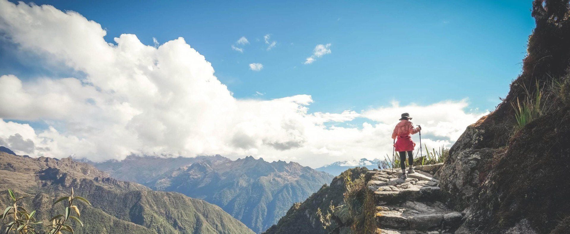 A person wearing a red jacket and black hat hikes up a stone path on a South American mountain, staying healthy while surrounded by dramatic clouds and rugged green peaks beneath a bright blue sky.