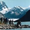 A group of people stands on a rocky shoreline by a lake, with snow-capped mountains and a glacier in the background under a clear blue sky—a breathtaking scene from an unforgettable South America tour.
