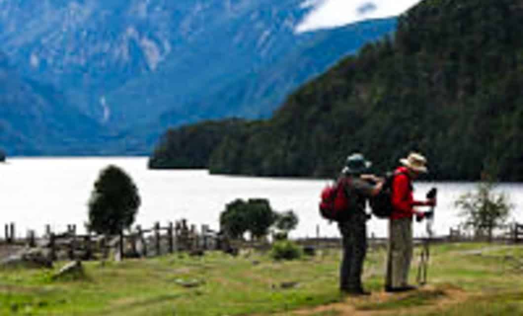 Two hikers with backpacks and walking sticks stand on grassy terrain near a wooden fence, overlooking a lake surrounded by mountains and forested hills near Mitico Puelo Lodge.