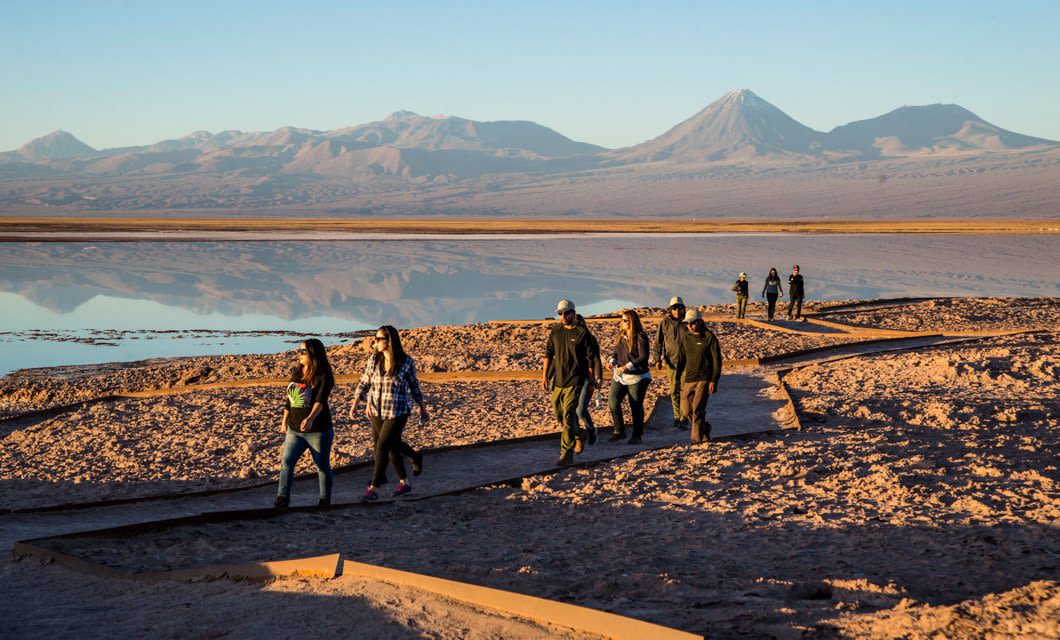 A group of people walk along a winding path by a reflective lake in the desert landscape near Cumbres Atacama Hotel, with distant mountains under a clear sky in the background.