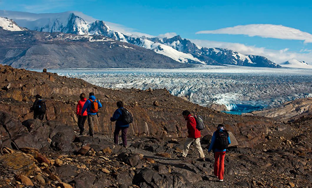 A group of hikers walk on rocky terrain with snow-capped mountains and a glacier in the background beneath a bright blue sky in Patagonia, Argentina.
