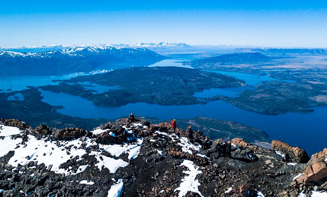 A panoramic view from a snowy mountain peak in Remota Patagonia, overlooking blue lakes, forested islands, and distant mountain ranges under a clear sky, with a few hikers visible on the rocky terrain.