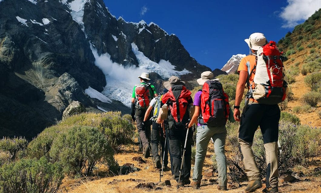 A group of hikers with backpacks and walking sticks trek up a rocky, grassy mountain trail toward snow-capped peaks under a clear blue sky, experiencing one of the top Bolivia activities for adventure seekers.