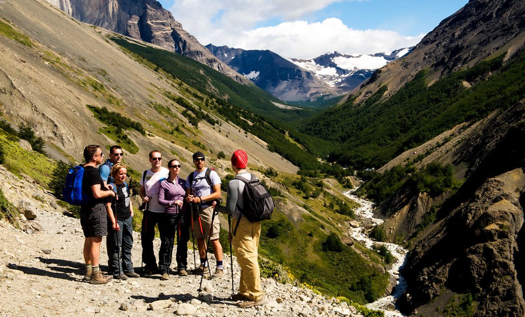 A group of hikers stands on a rocky mountain trail in Patagonia, talking and looking at the scenic valley below, surrounded by green slopes, rugged cliffs, and distant snow-capped peaks under a partly cloudy sky.
