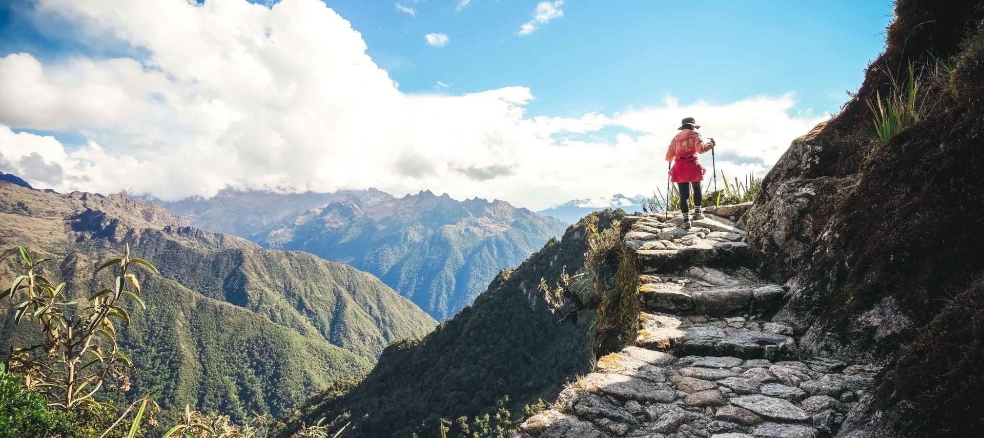 A person in hiking gear walks along a stone path on a mountain trail at Machu Picchu, surrounded by lush green peaks and dramatic clouds under a bright blue sky.