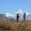 Quick note: Two hikers with backpacks stand on a grassy hilltop, with snow-capped mountains and a clear blue sky in the background.