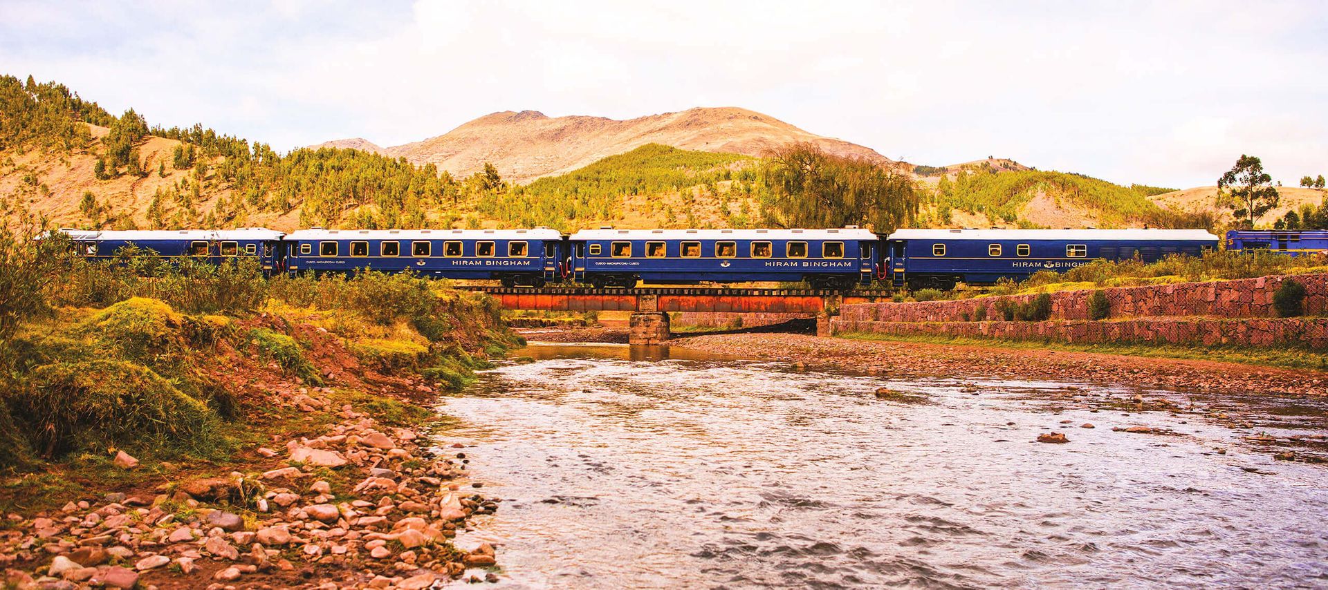 A blue passenger train, part of Peru Train Tours, crosses a small bridge over a shallow river, with green hills and mountains in the background under a partly cloudy sky.