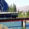 A train crosses a red bridge over a blue river, with trees and rocky mountains in the background under a clear sky.