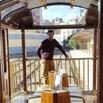 A person stands on the deck of a wooden boat with a glass roof, holding onto the railing. Buildings and greenery are visible in the background under a blue sky.