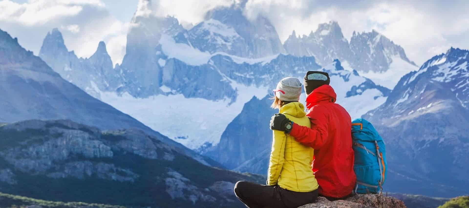 Two hikers in bright jackets sit on a rock, embracing as they admire a dramatic South America mountain landscape with snowy peaks and clouds—a picture-perfect moment for Honeymoon Tours.