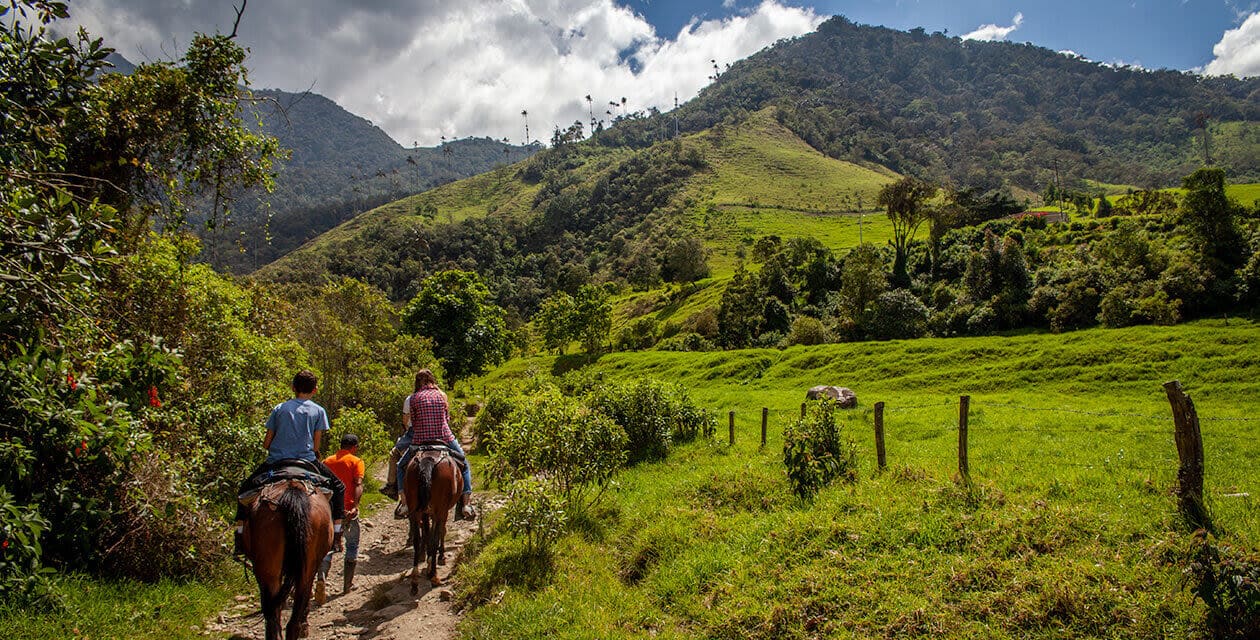 Three people riding horses along a dirt path through lush green fields and hills, offered by Colombia Adventure Tours, with trees and a partly cloudy sky in the background.