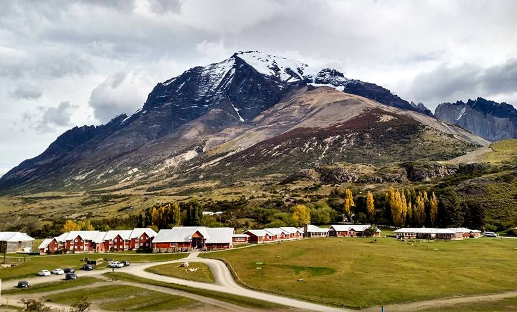 A row of red buildings with gray roofs, part of Hotel Las Torres, stands on a grassy plain with parked cars nearby. In the background, a towering, snow-capped mountain rises under a cloudy sky.