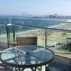 A glass table and wicker chair on a balcony promise a nice experience, overlooking a sandy beach with many people, blue ocean waves, and a distant cityscape under a clear sky.