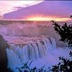 A wide waterfall cascades over rocky cliffs, surrounded by mist and lush greenery, with a well-arranged scene set against a colorful sunset sky, creating an unforgettable experience.
