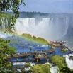 A scenic view of Iguazu Falls with lush greenery, mist rising from the cascading waterfalls, a rainbow, and people on a walkway—perfect for your itinerary with any top-rated tour company.