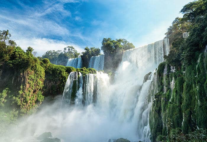 Powerful waterfalls cascade over lush, green cliffs surrounded by dense vegetation—a must-see among Argentina attractions—with mist rising from the water and a bright blue sky overhead.
