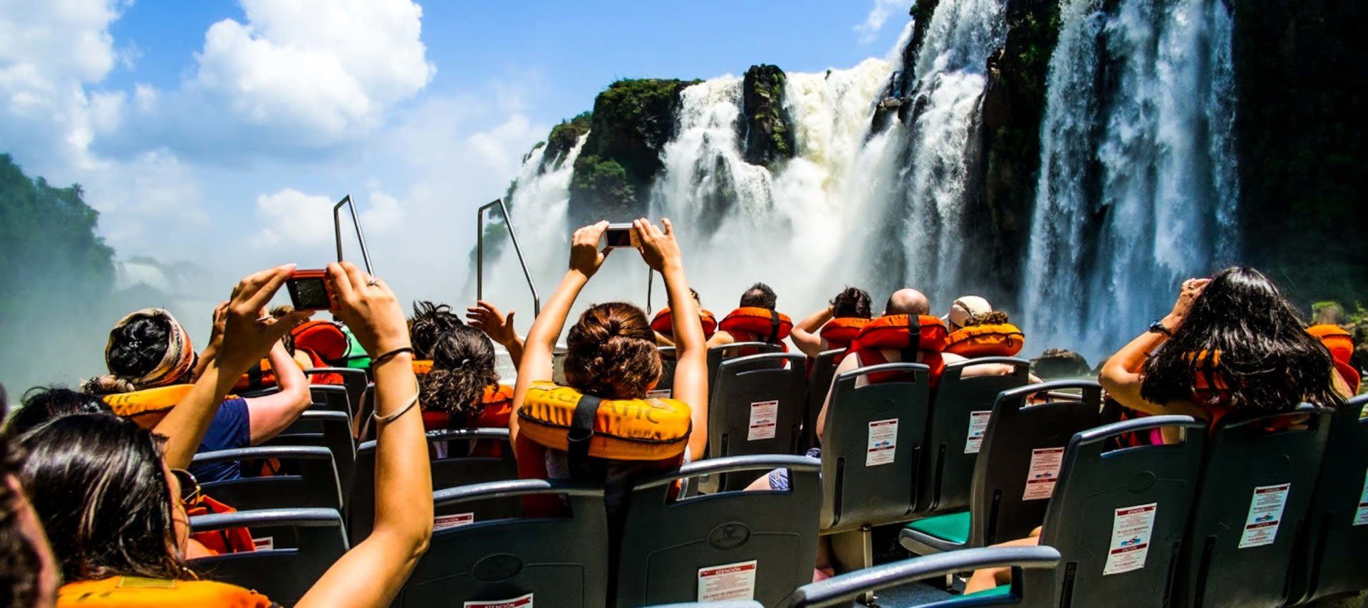 A group of people wearing life jackets take photos from a boat as they approach the powerful Iguazu Falls under a bright blue sky with scattered clouds, enjoying one of the thrilling Iguazu Falls tours.