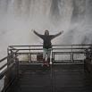 A person in a hooded jacket stands on a metal platform with arms raised, facing the amazing, misty Iguassu waterfall in the background.