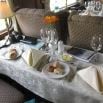 A small dining table set for two with white napkins, wine glasses, bread rolls, and a plate of pastries evokes the cozy charm of cafes in Bolivia. The lace tablecloth and yellow flowers by the window add a warm, inviting touch.