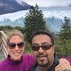 A smiling couple takes a selfie outdoors with trees behind them, a large glacier, and mountains in the background under a cloudy sky on their South American honeymoon.