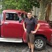 A person stands in front of a vintage red pickup truck, smoothly posing with one hand on their hip and the other on the truck’s open door, as if ready for a trip near trees and a building.