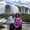 A man and woman stand together in front of a railing with large, cascading waterfalls and lush greenery in the background, enjoying the sights on a well organized tour under a partly cloudy sky.